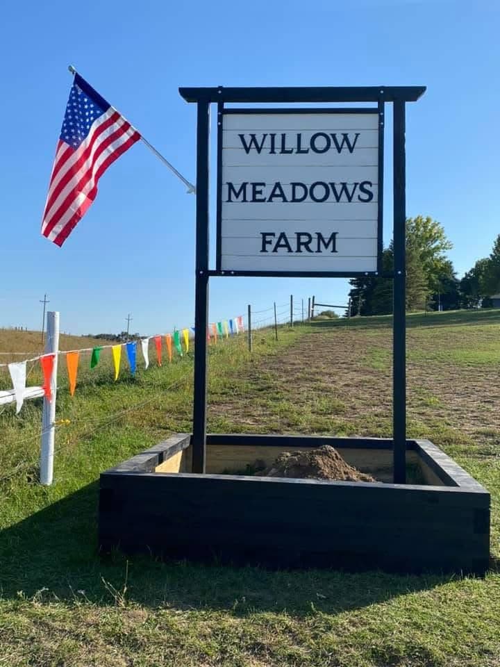 The Willow Meadows Farm roadside sign with an American flag and colorful pennant flags at the farm entrance on Hwy 115